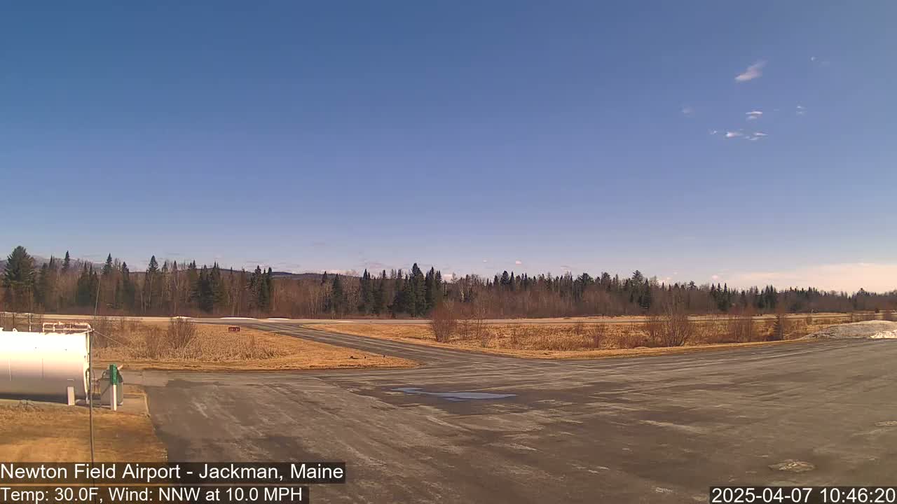 An airfield with a large, mostly empty tarmac area, a white fuel tank, and a line of trees in the background under a clear blue sky.