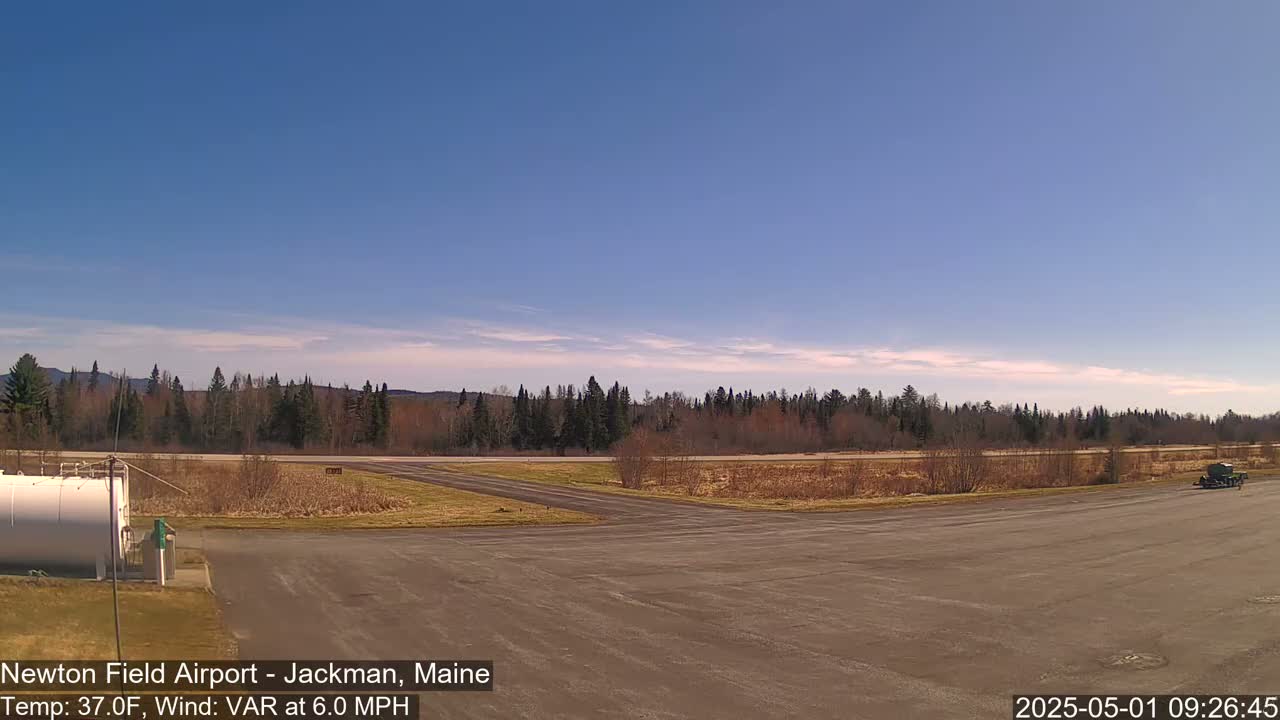 A mostly clear blue sky over a flat, empty airfield bordered by a line of trees and a distant mountain range under a light wind.
