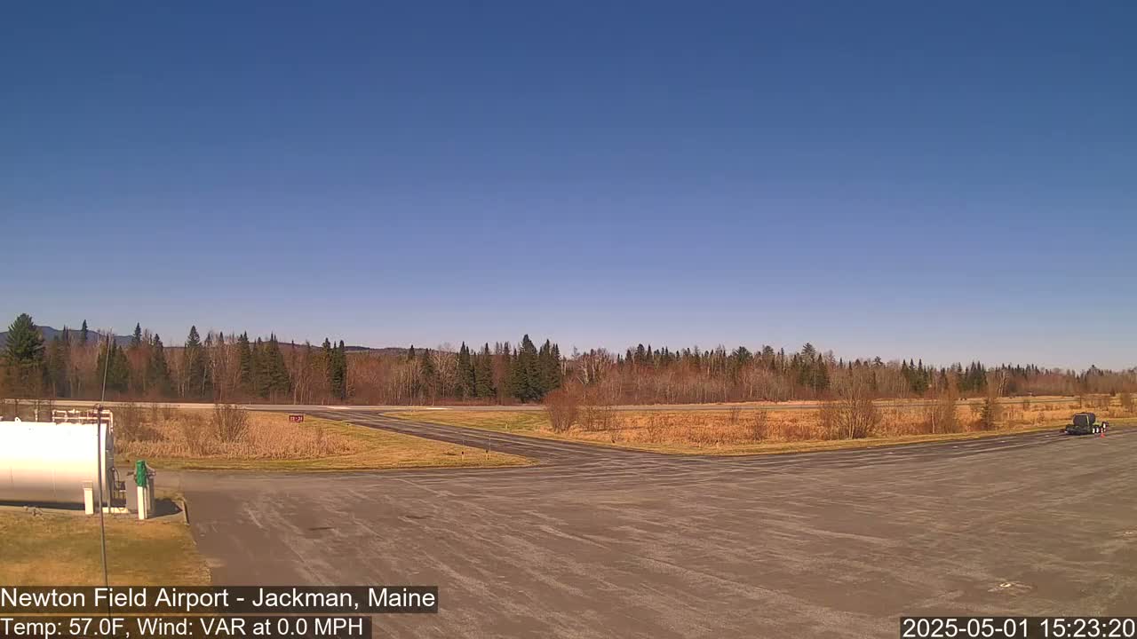 A mostly clear, sunny day reveals an airfield with a large expanse of tarmac, surrounded by a line of trees and a grassy field.