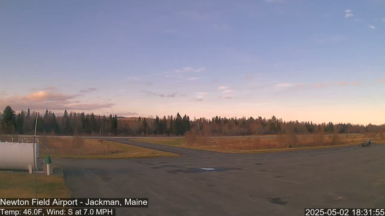 A mostly clear, twilight sky hangs over a small airport with a paved runway and a line of trees in the background, under a light south wind.