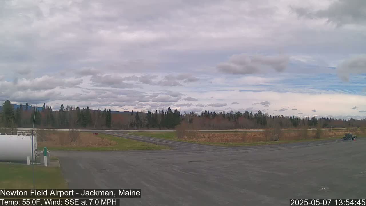 A partly cloudy sky over a flat, paved airfield with a line of trees and a vehicle in the distance.