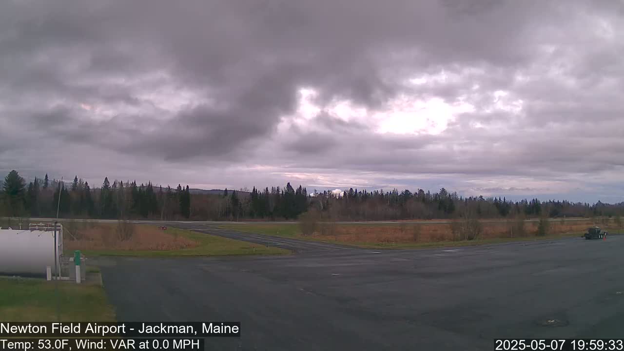 A mostly cloudy sky hangs over an airfield with a paved runway and grassy areas, bordered by a line of trees.