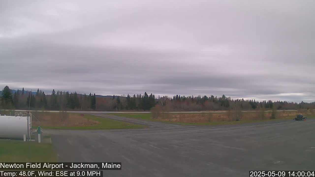 A mostly overcast sky hangs over an airfield, where a vehicle is parked near a large, white tank, and a line of trees stretches across the horizon.
