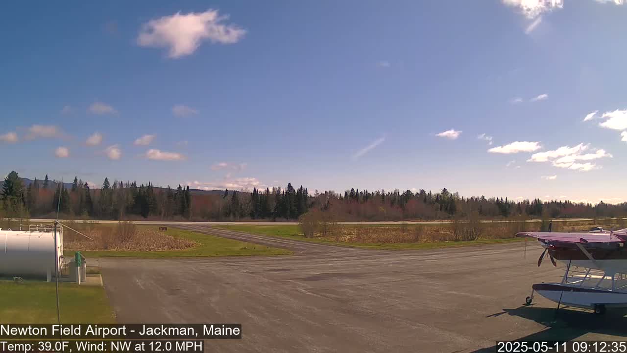 A small airport with a seaplane parked on the tarmac is seen under a partly cloudy sky with a northwest wind.