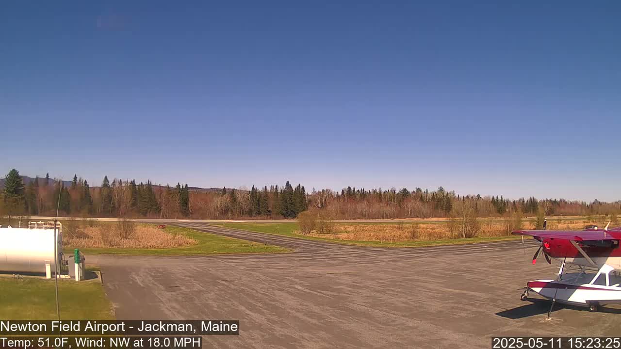 A small airfield with a red and white seaplane parked on the tarmac, under a clear blue sky, with a light wind and trees in the background.
