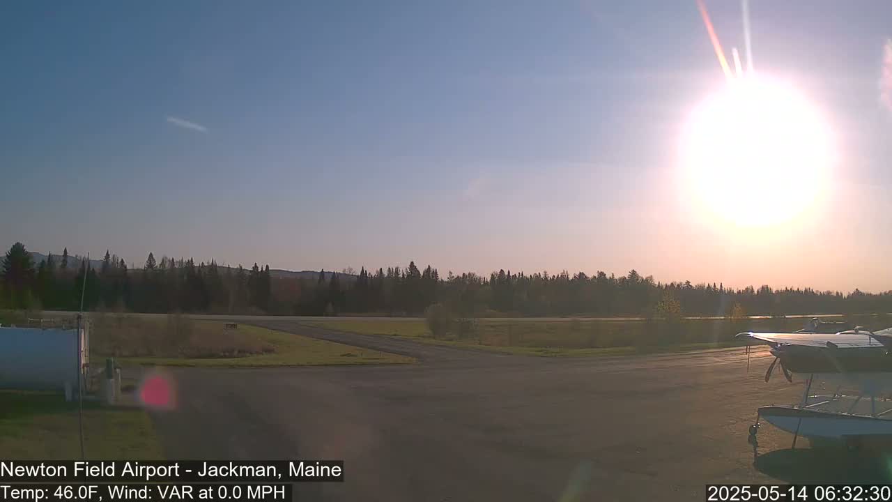 A bright sun shines over a calm airfield with a seaplane parked,  backed by a line of trees under a clear sky.