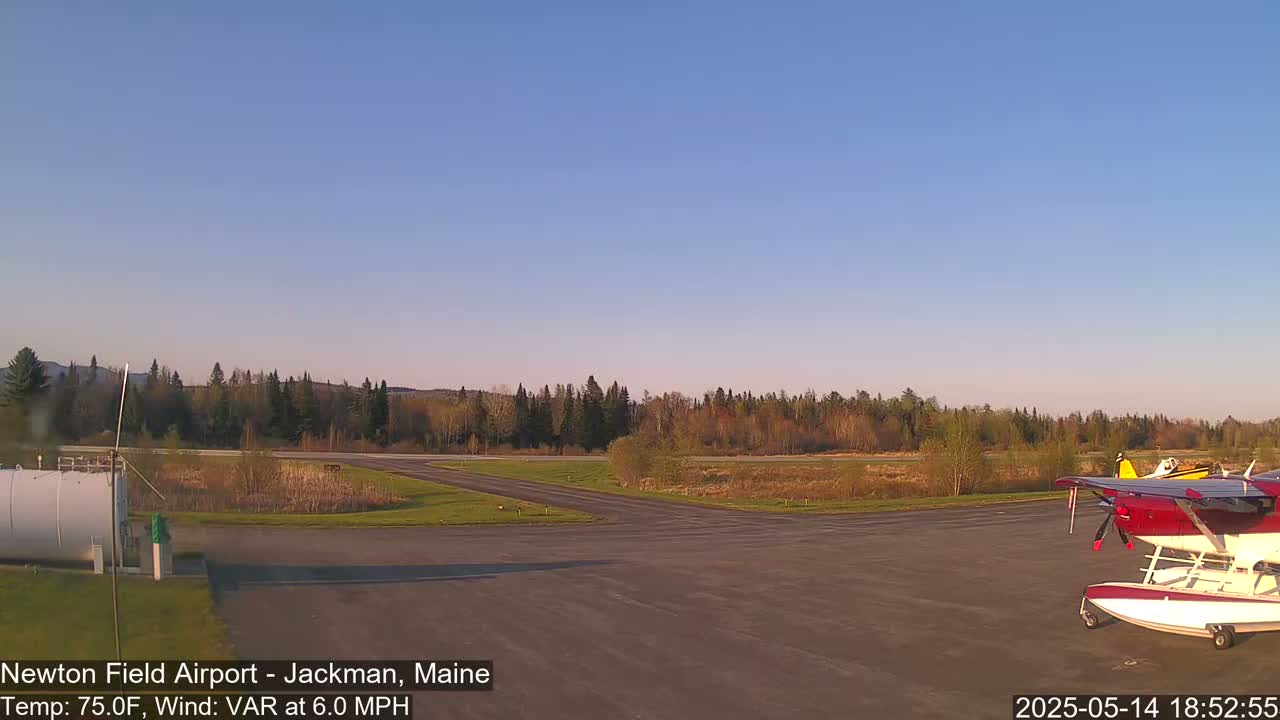Under a clear, blue sky, a small airport shows a red and white seaplane parked on the tarmac, surrounded by green grass and trees, with a calm wind.