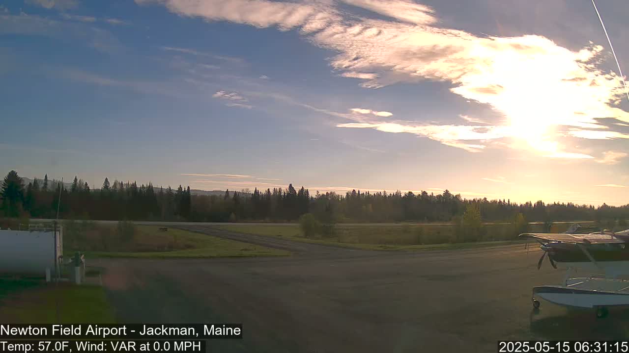 A small airplane sits on a tarmac at sunrise under a partly cloudy sky, with a line of trees visible in the background.