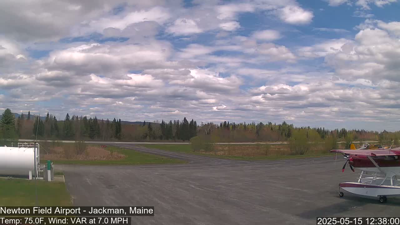 A partly cloudy sky over a small airport with a seaplane parked on the tarmac and a line of trees in the background.