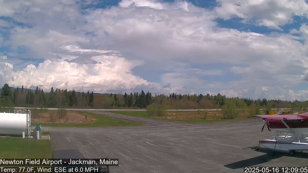 A partly cloudy sky over an airport tarmac with a seaplane parked and a grassy area with trees in the background.