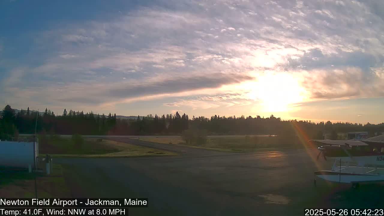 A sunrise illuminates a partly cloudy sky over a small airport with a seaplane parked on the tarmac, next to a line of trees.