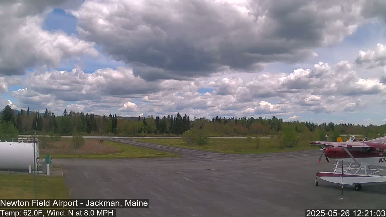 A seaplane sits on a tarmac at an airport under a partly cloudy sky, with a line of trees visible in the background.