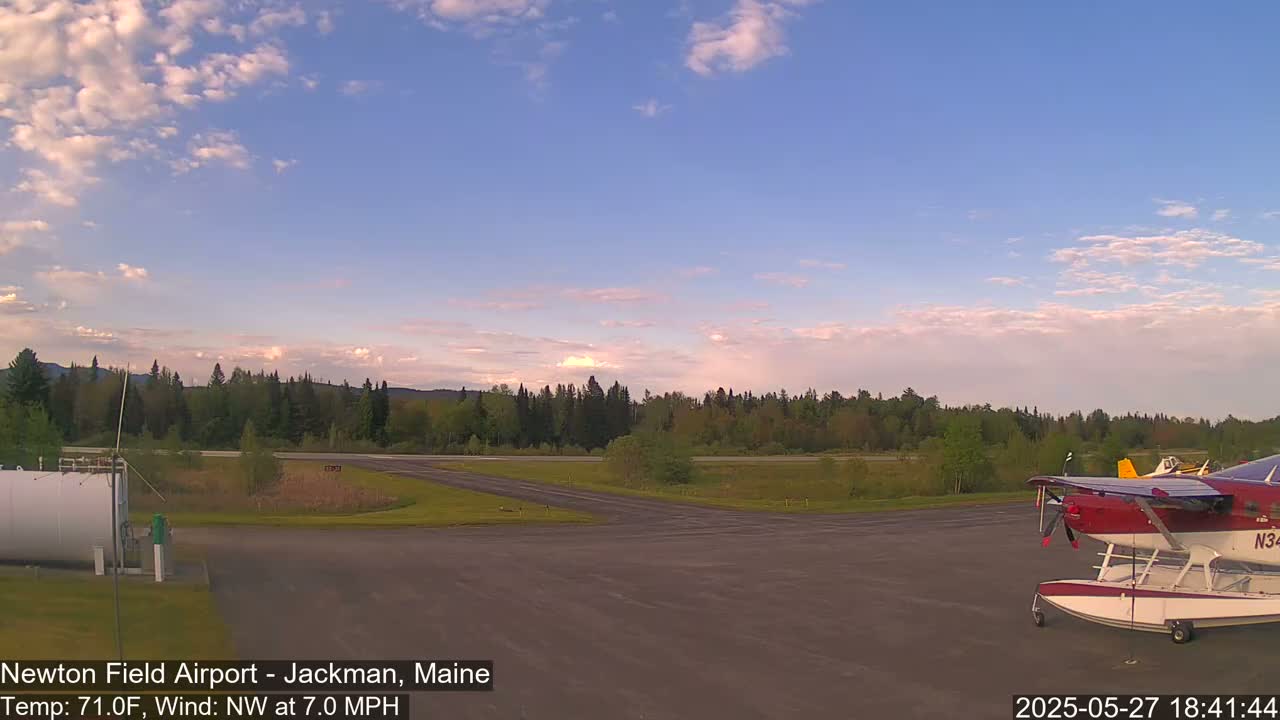 A seaplane is parked on an airport tarmac under a partly cloudy sky at sunset, with a line of trees in the background.