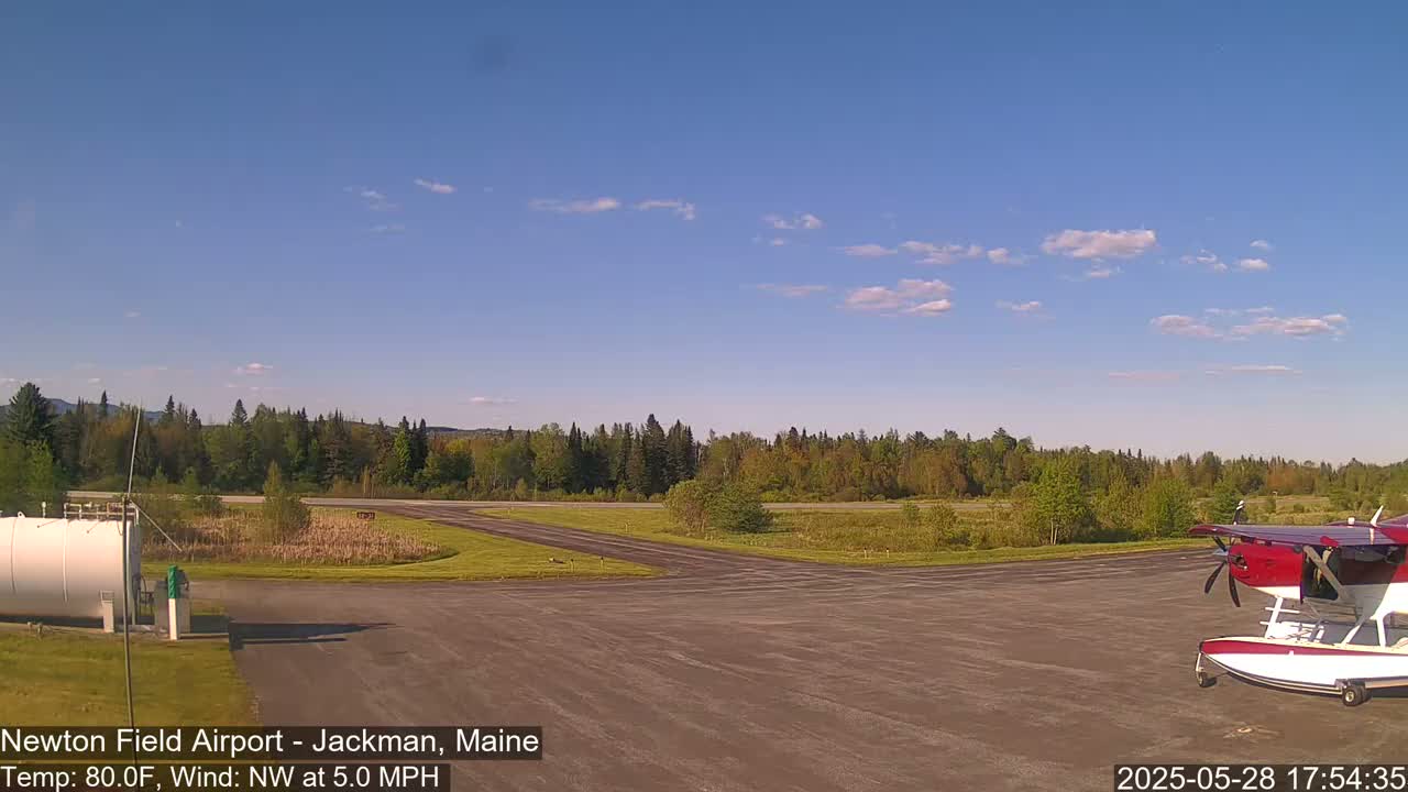 A small red and white seaplane is parked on an airfield surrounded by trees under a mostly clear, blue sky.