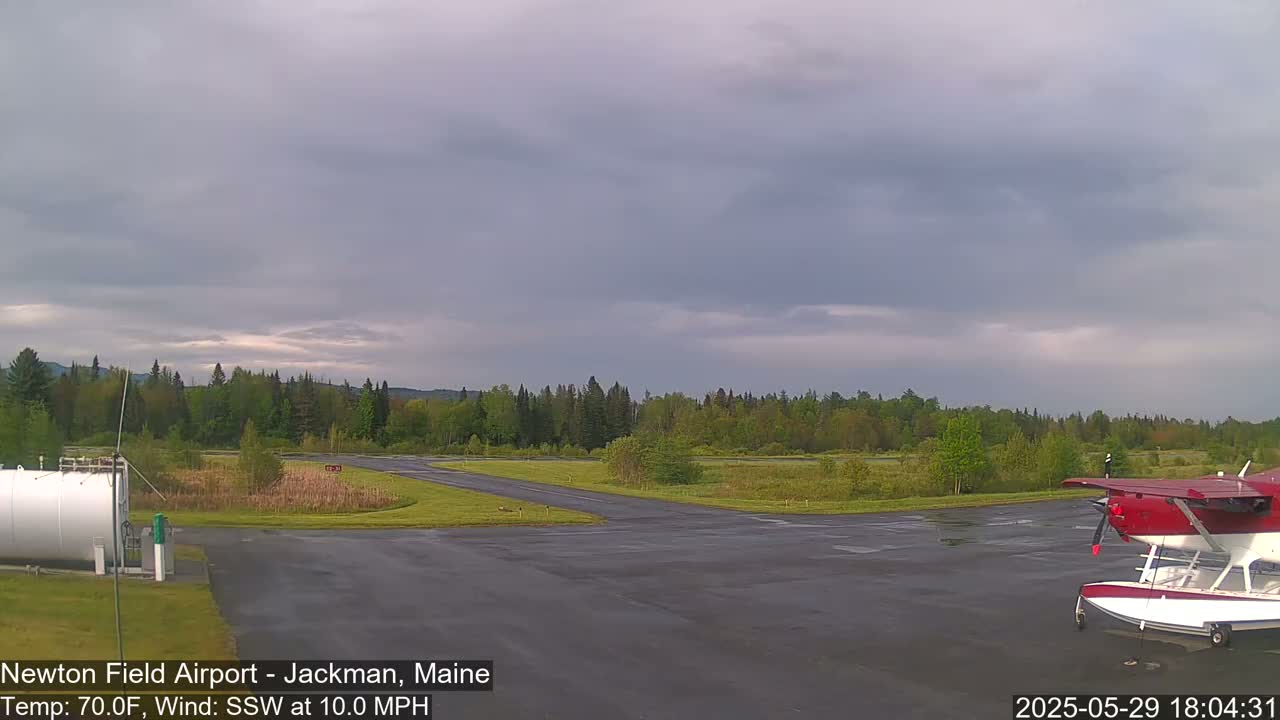 A red seaplane sits on a tarmac next to a grassy airfield under a mostly cloudy sky.