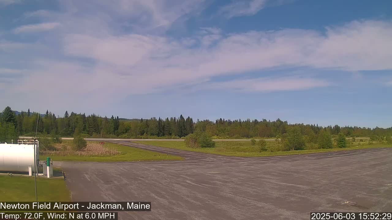 A mostly sunny day reveals a small airport runway bordered by green trees and a grassy field under a partly cloudy sky.