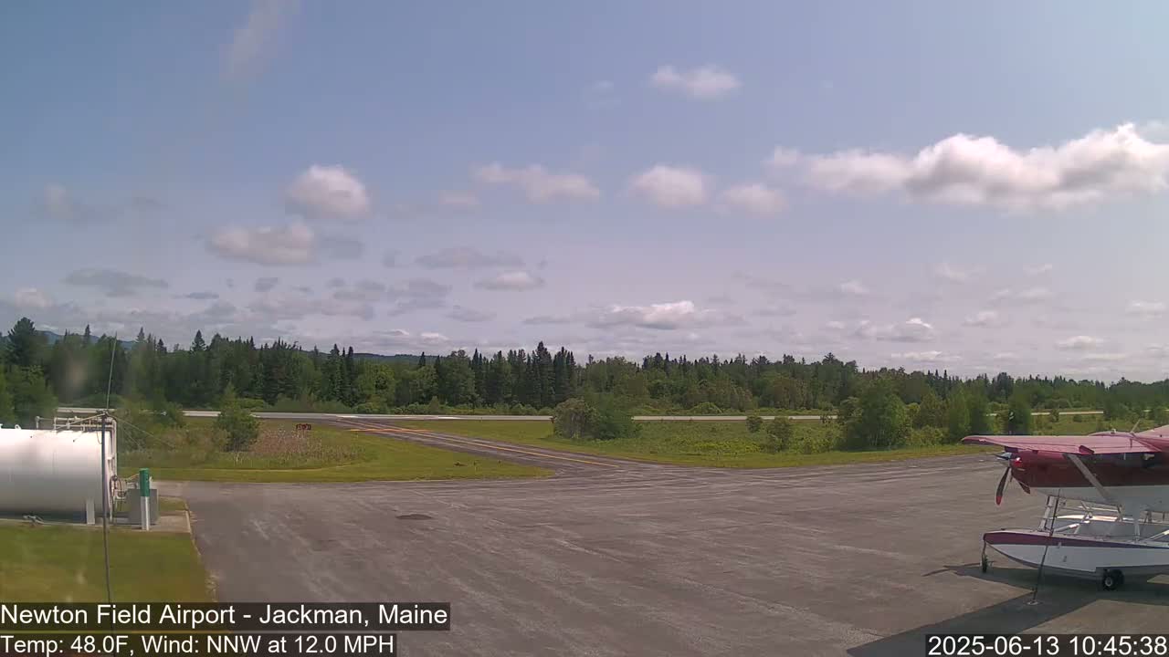 A small seaplane is parked on an airport tarmac, with a line of trees and a partly cloudy sky in the background.