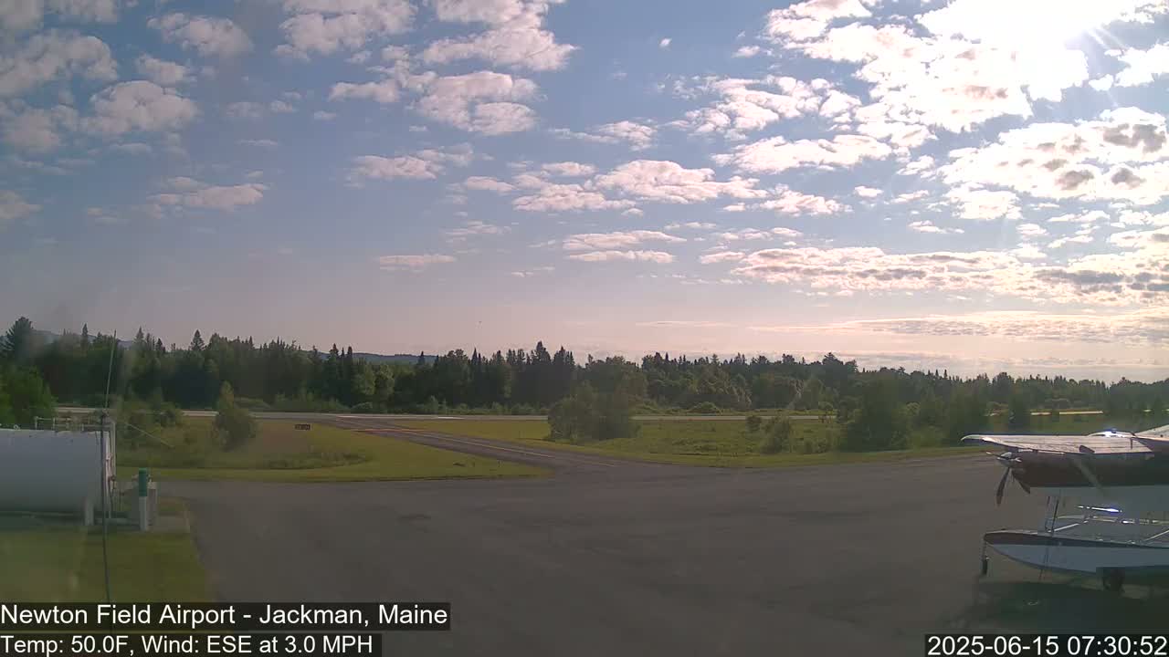 A partly cloudy sky over a small airport with a seaplane partially visible on the tarmac and a line of trees in the distance.