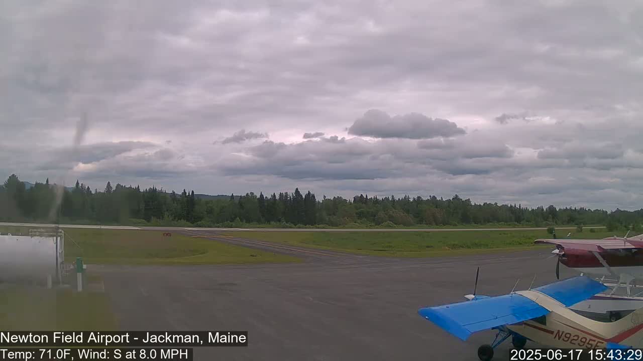 A partly cloudy sky over an airport with two small airplanes parked on the tarmac and a line of trees in the background.