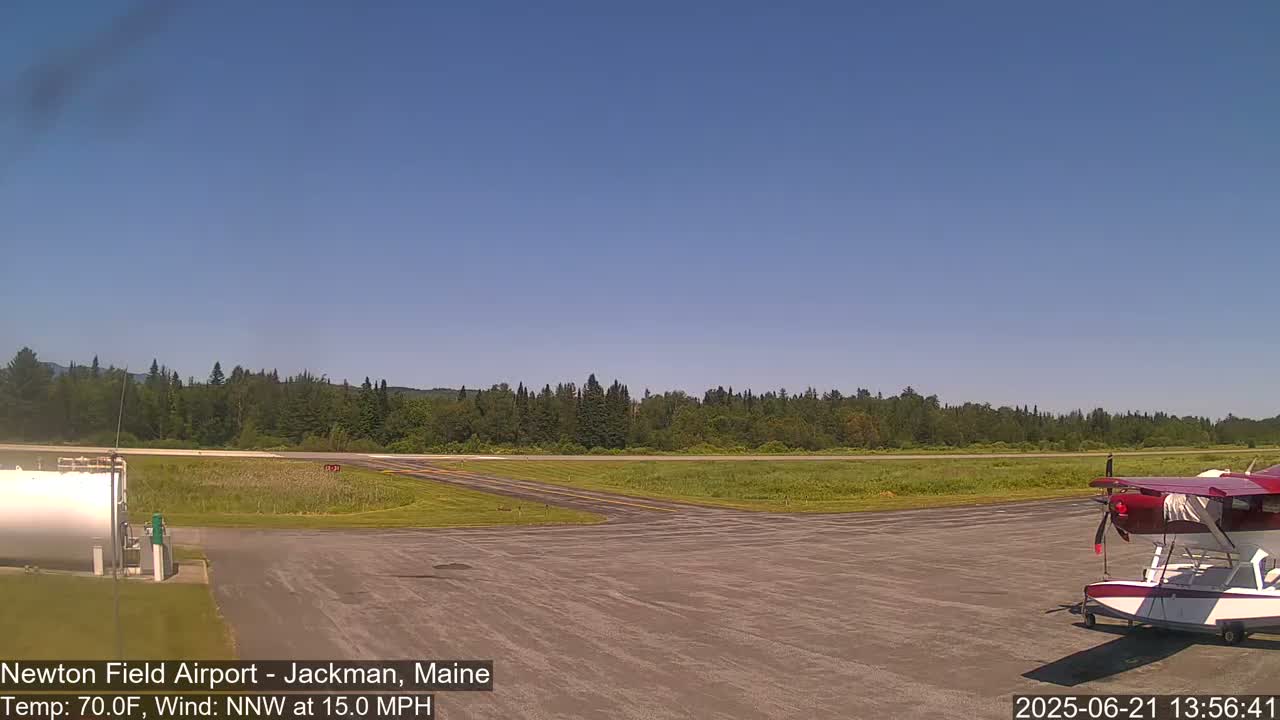 A small red seaplane sits on a tarmac at an airfield on a sunny day with a north-northwest wind of 15 mph.