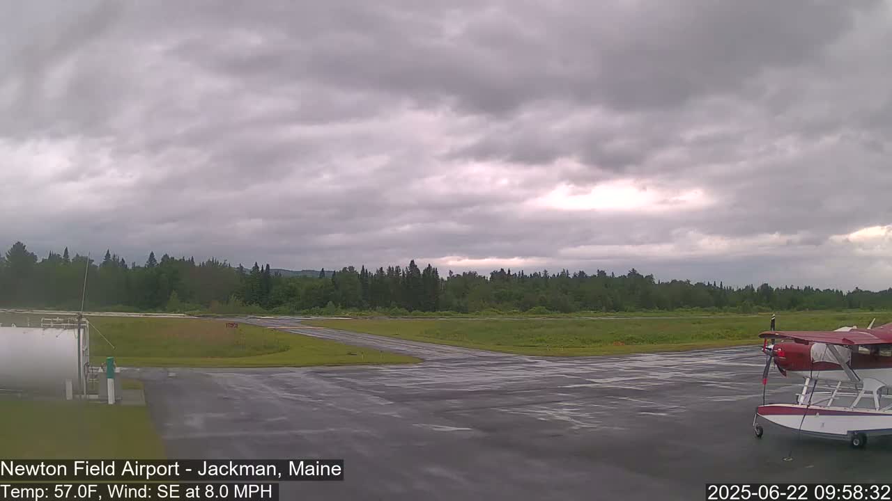 A red seaplane sits on a wet tarmac at a small airport under an overcast sky.