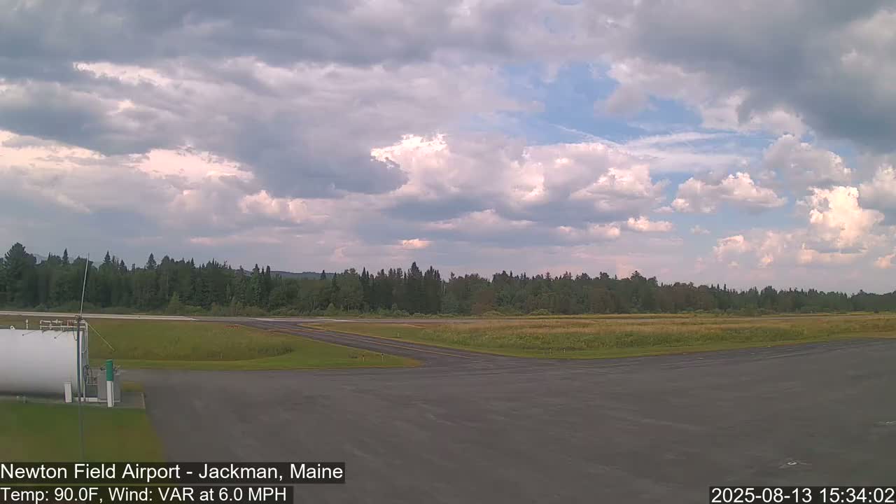 A partly cloudy sky over a flat airfield with a grassy field and a line of trees in the background, under warm conditions with a light, variable wind.