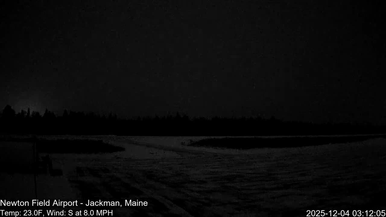 A vast, snow-covered landscape at night extends towards a distant, dark tree line under an almost completely black sky, indicating cold and extremely low light conditions.