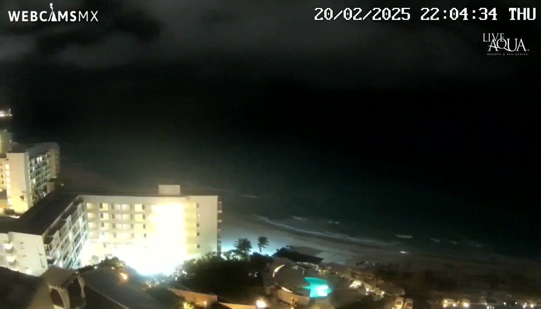 A nighttime view shows a beach with gently lapping waves under a dark, cloudy sky, and brightly lit hotels and a swimming pool are visible in the foreground.