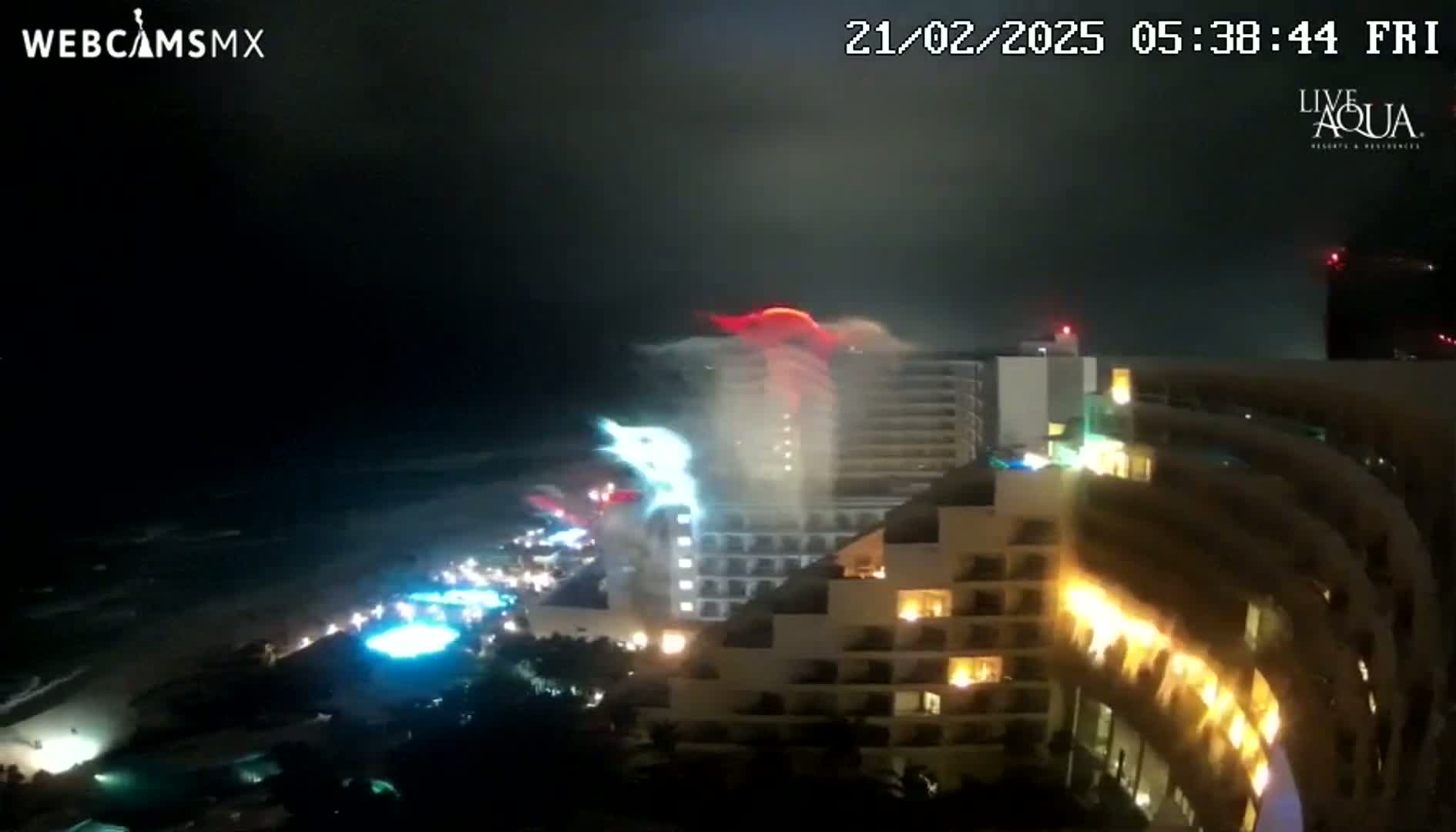 A nighttime view of a coastal city shows a large hotel with lights on and fireworks going off near the ocean under a dark sky.