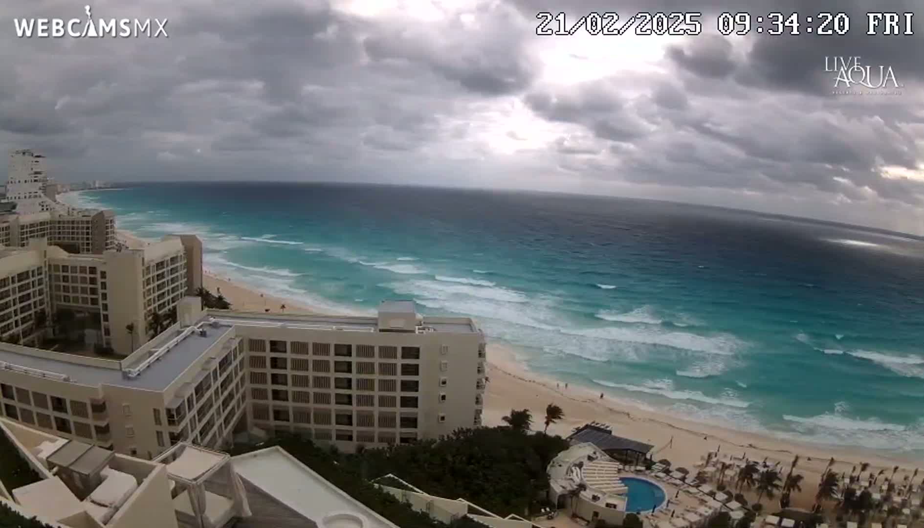 An ocean beach with turquoise water and moderate waves is seen from an elevated viewpoint overlooking several multi-story hotels under a cloudy sky.