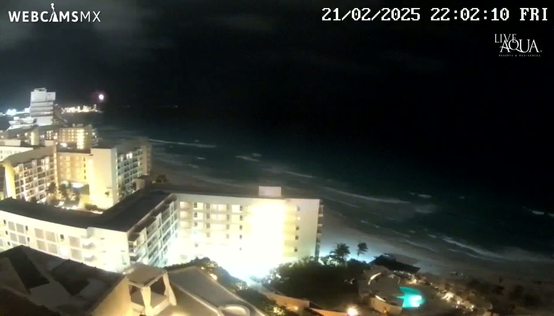 A nighttime aerial view shows a beach with gently rolling waves, illuminated beachfront hotels, and a swimming pool.