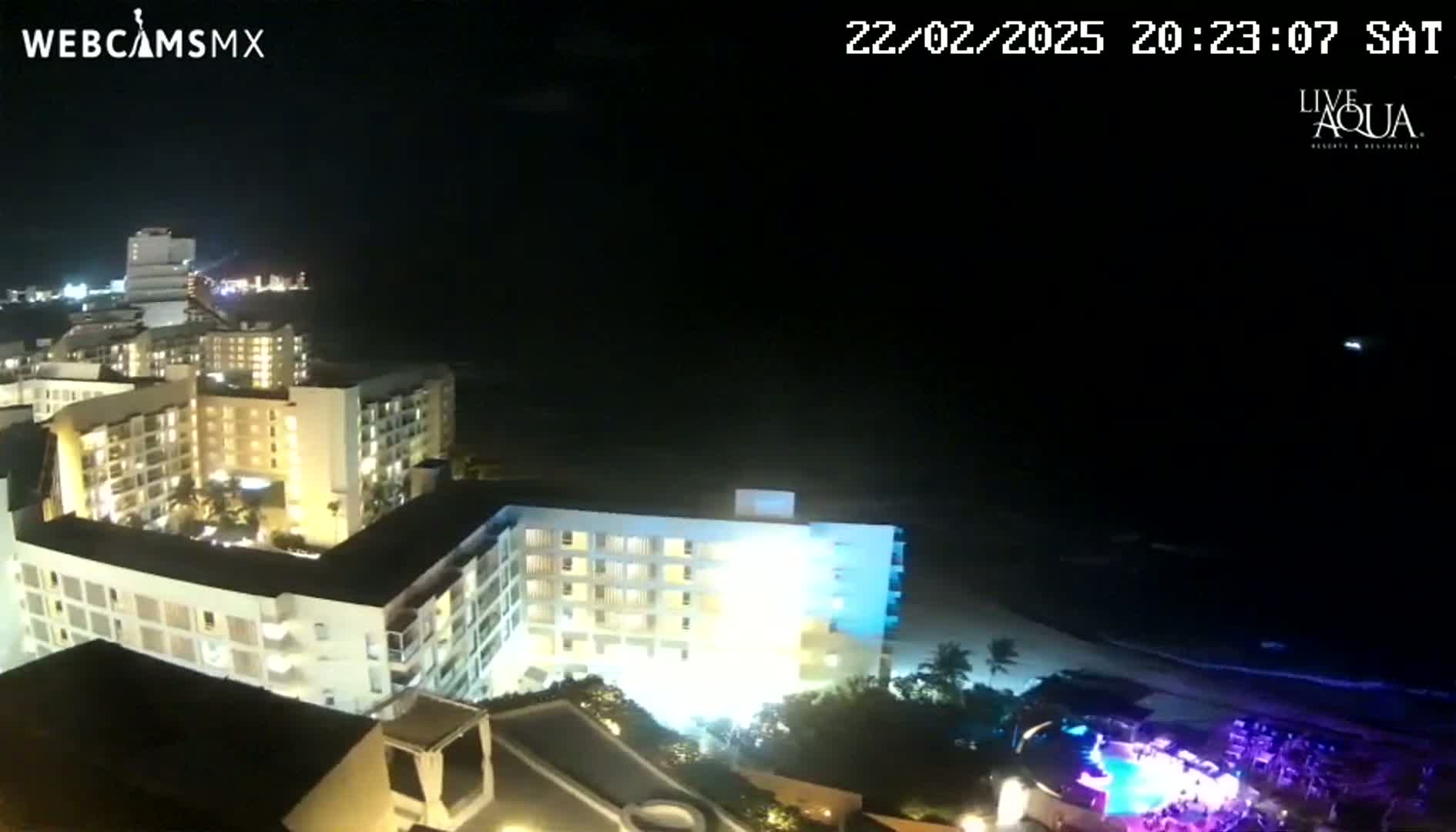 A nighttime, aerial view shows brightly lit resort buildings near a dark ocean under a clear sky.