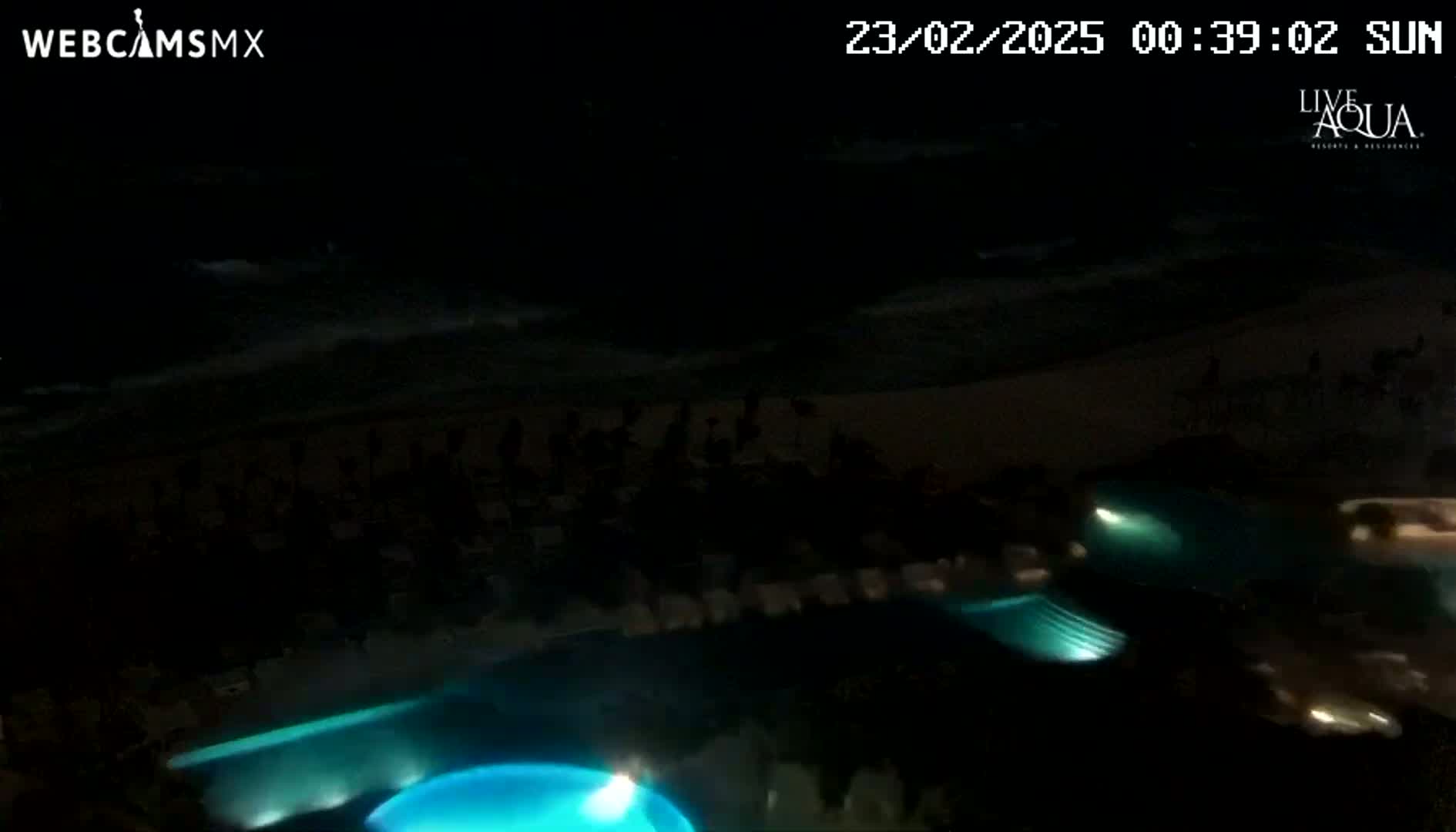 A nighttime view shows a dark beach and ocean with several illuminated swimming pools at a resort.