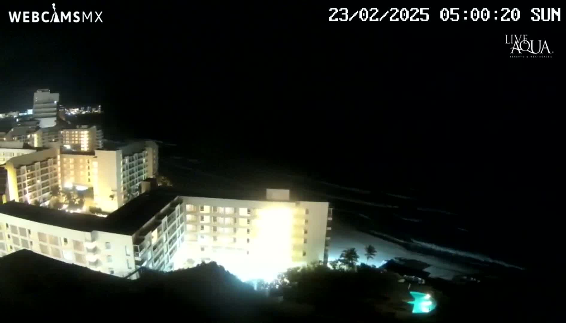 A nighttime view of brightly lit multi-story buildings near a dark ocean under a clear, dark sky.