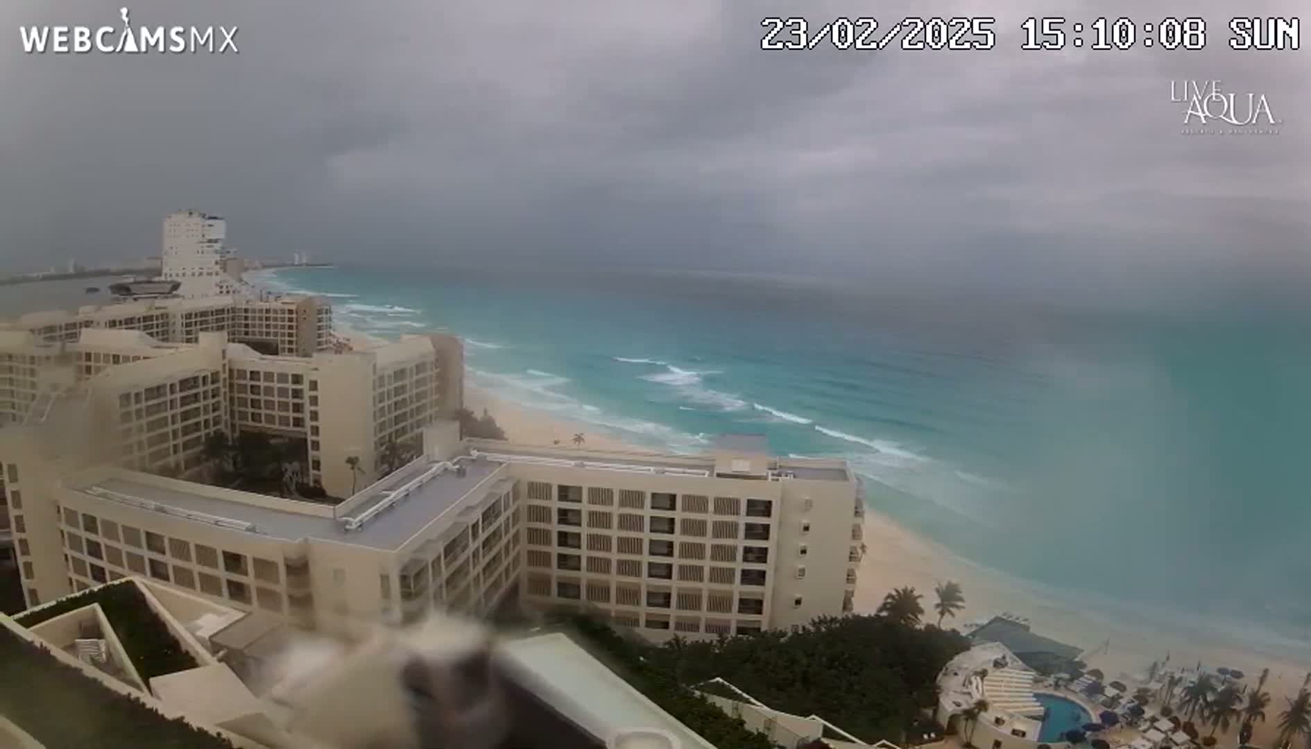 An overcast sky hangs over a beach and a large resort complex, with turquoise ocean waves breaking on the shore.