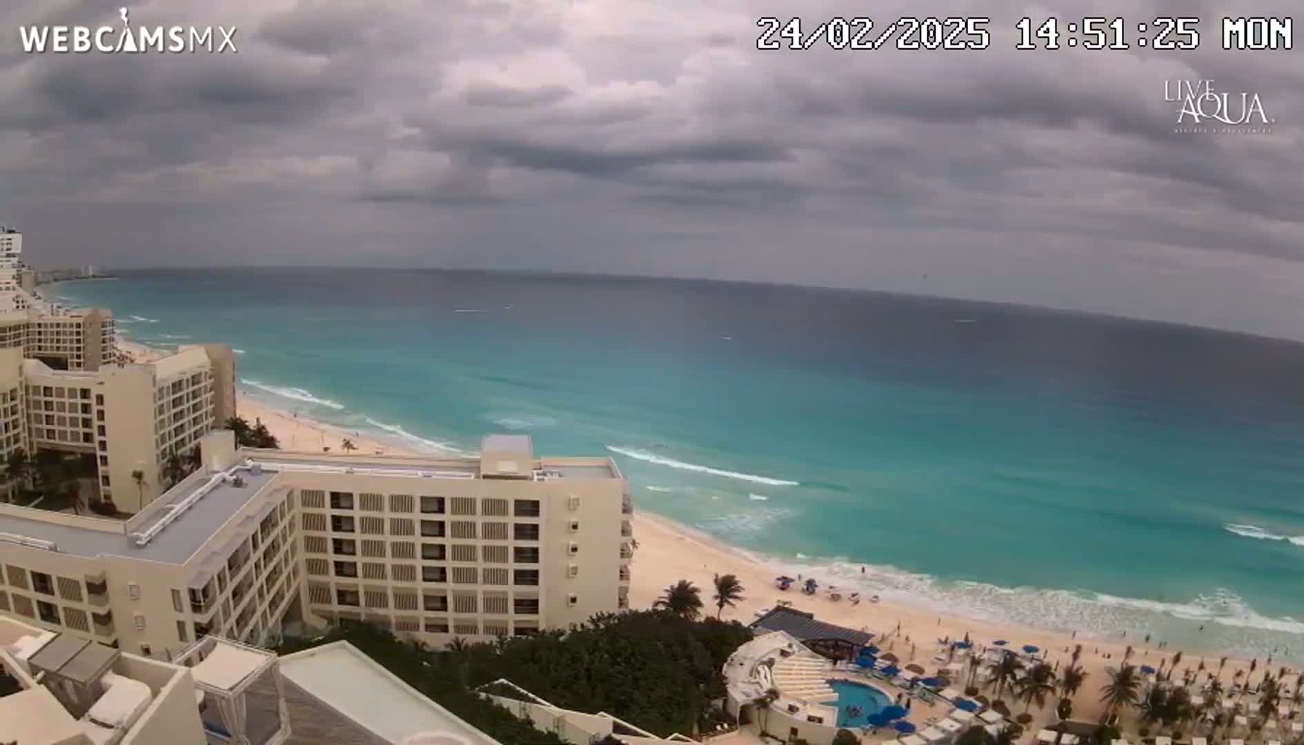 An elevated view shows a beach with turquoise water and several high-rise hotels under a cloudy sky.