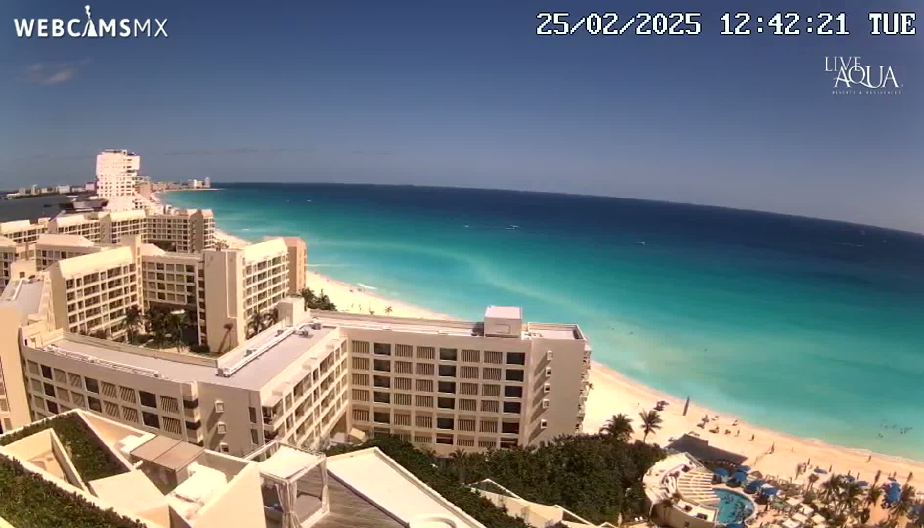 A high-angle view shows several large beige hotels situated near a beach with clear, turquoise water under a bright, sunny sky.