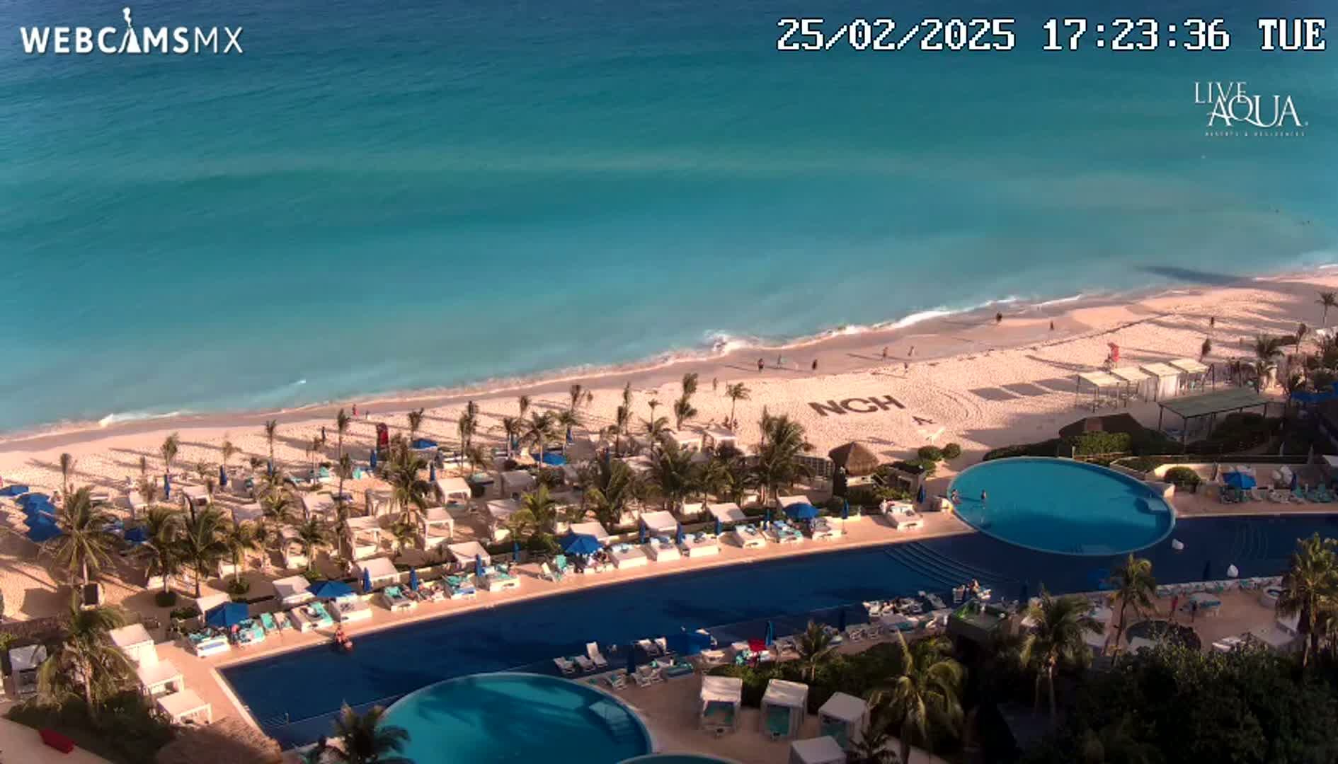 An aerial view shows a resort with a large, curved pool next to a beach with turquoise water, under sunny skies.