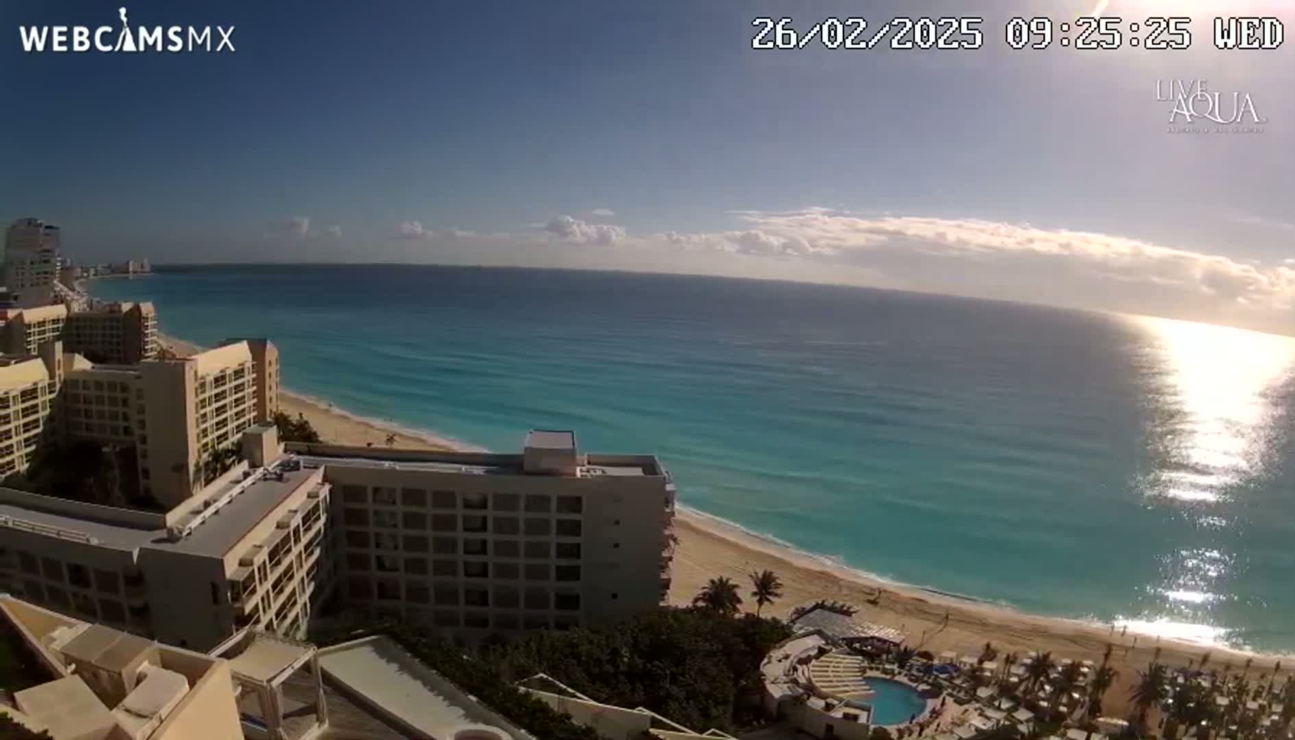 An elevated view shows a sunny beach with turquoise water and several high-rise hotels under a mostly clear sky.
