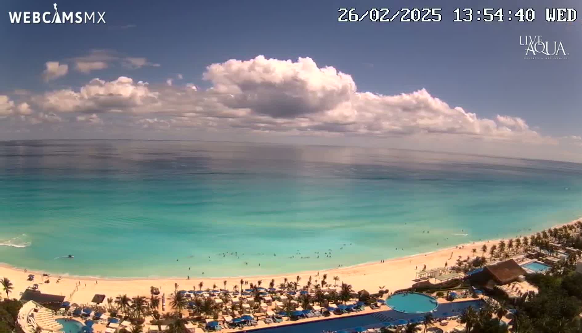 A partly cloudy sky overlooks a tropical beach with turquoise water, palm trees, and a resort with a pool.