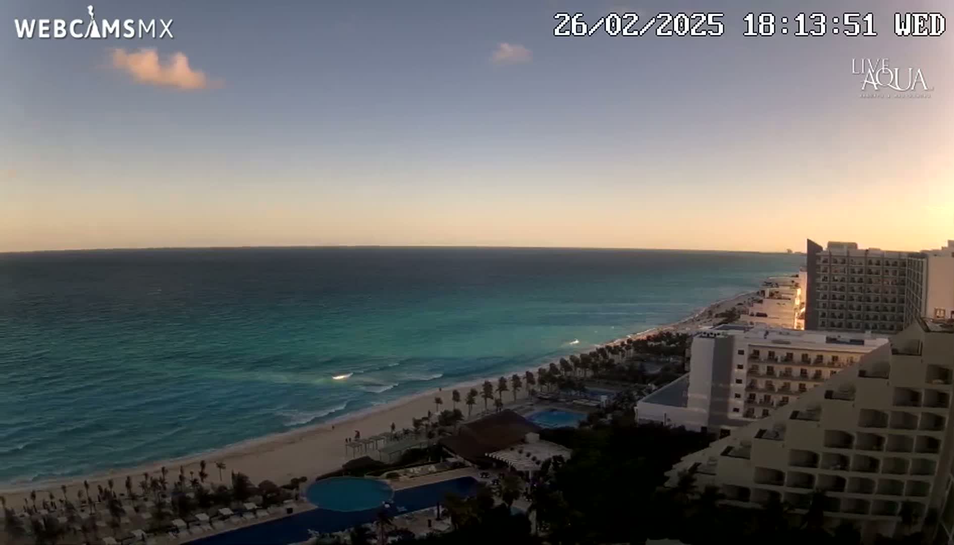 An ocean beach at sunset with calm waves, a sandy shoreline, and several large hotels visible in the foreground.
