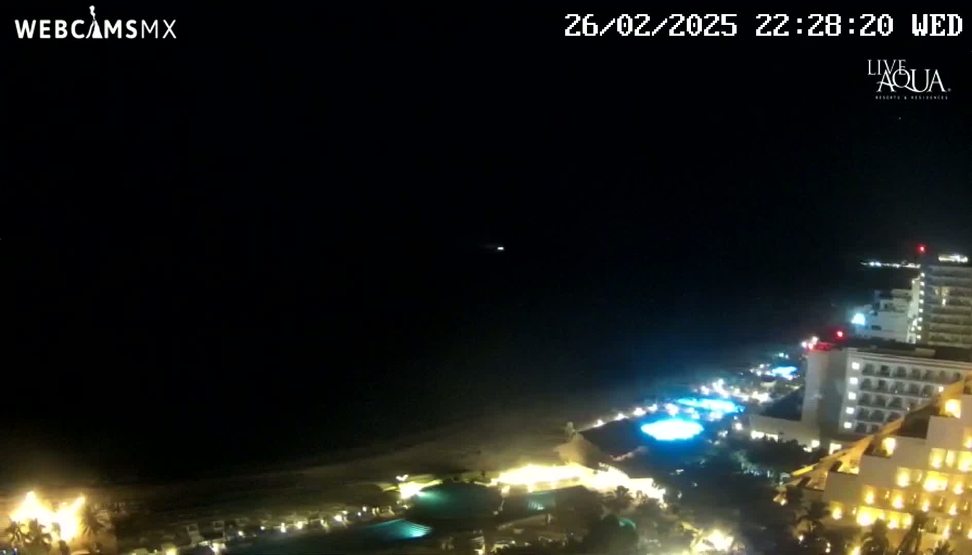 A nighttime, high-angle view shows a coast with illuminated resort buildings and pools along a dark beach under a clear, dark sky.