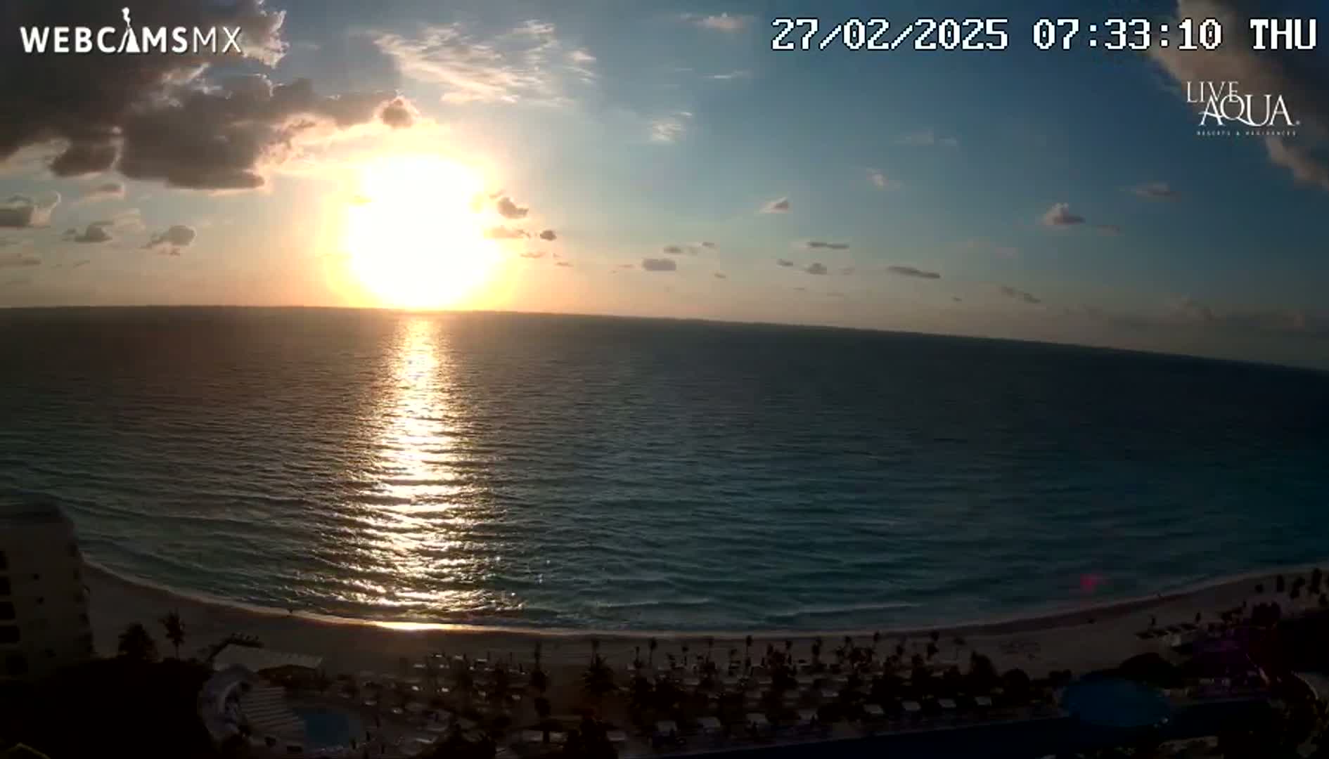 A sunrise over a calm ocean with a sandy beach and resort visible in the foreground under partly cloudy skies.
