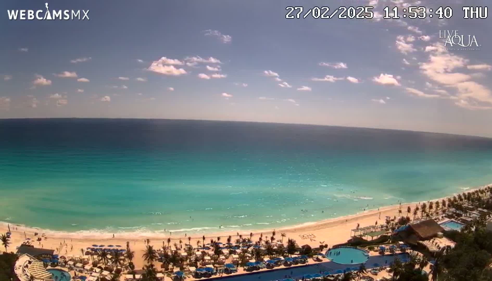 A mostly sunny day shows a turquoise ocean meeting a white sand beach lined with palm trees and resort amenities.