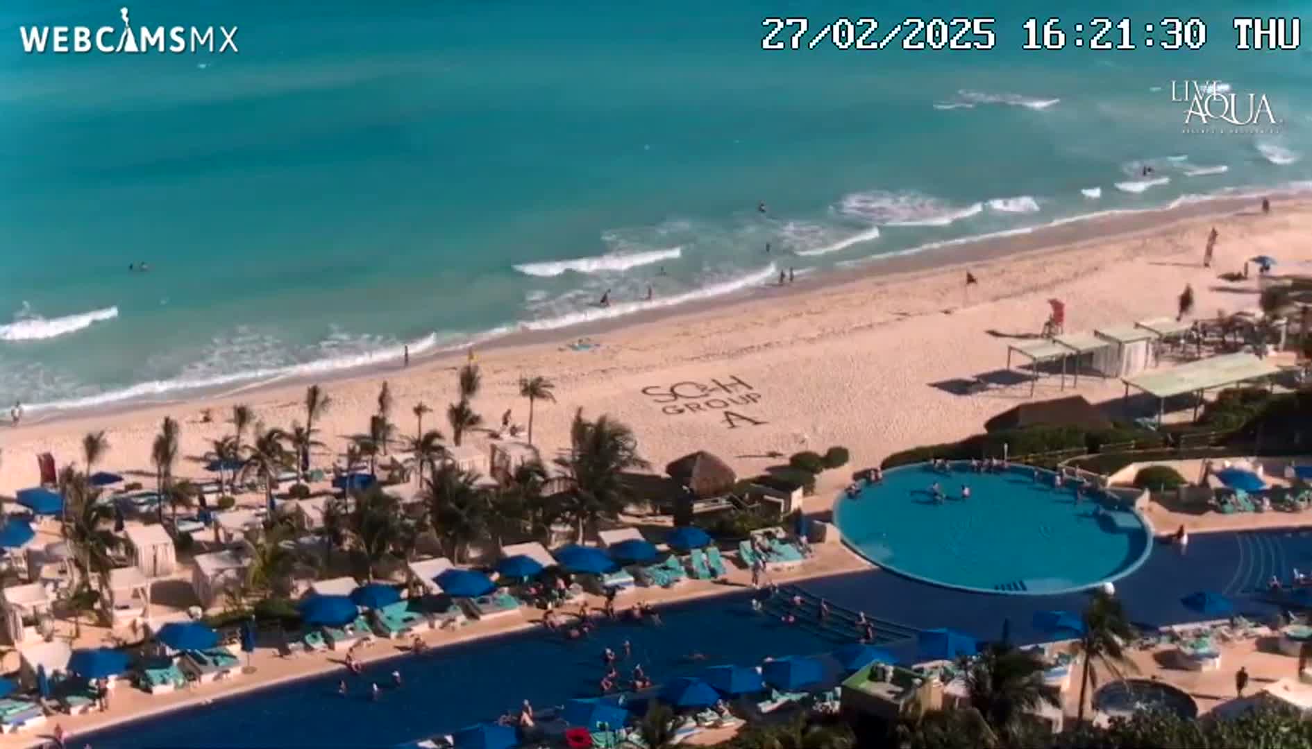 An aerial view shows a beach with turquoise water and gentle waves, a resort pool with numerous blue umbrellas and lounge chairs, and palm trees surrounding the area on a sunny day.