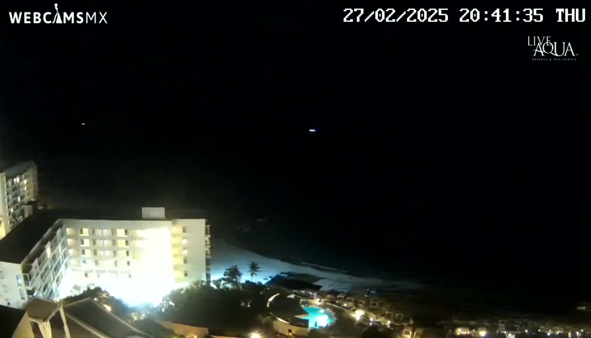 A nighttime view of a brightly lit resort complex next to a dark ocean beach under a clear, dark sky.