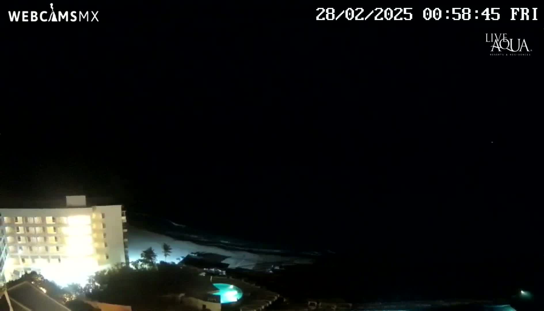 A brightly lit beachfront building is shown at night, overlooking a dark ocean with small waves under a clear, dark sky.