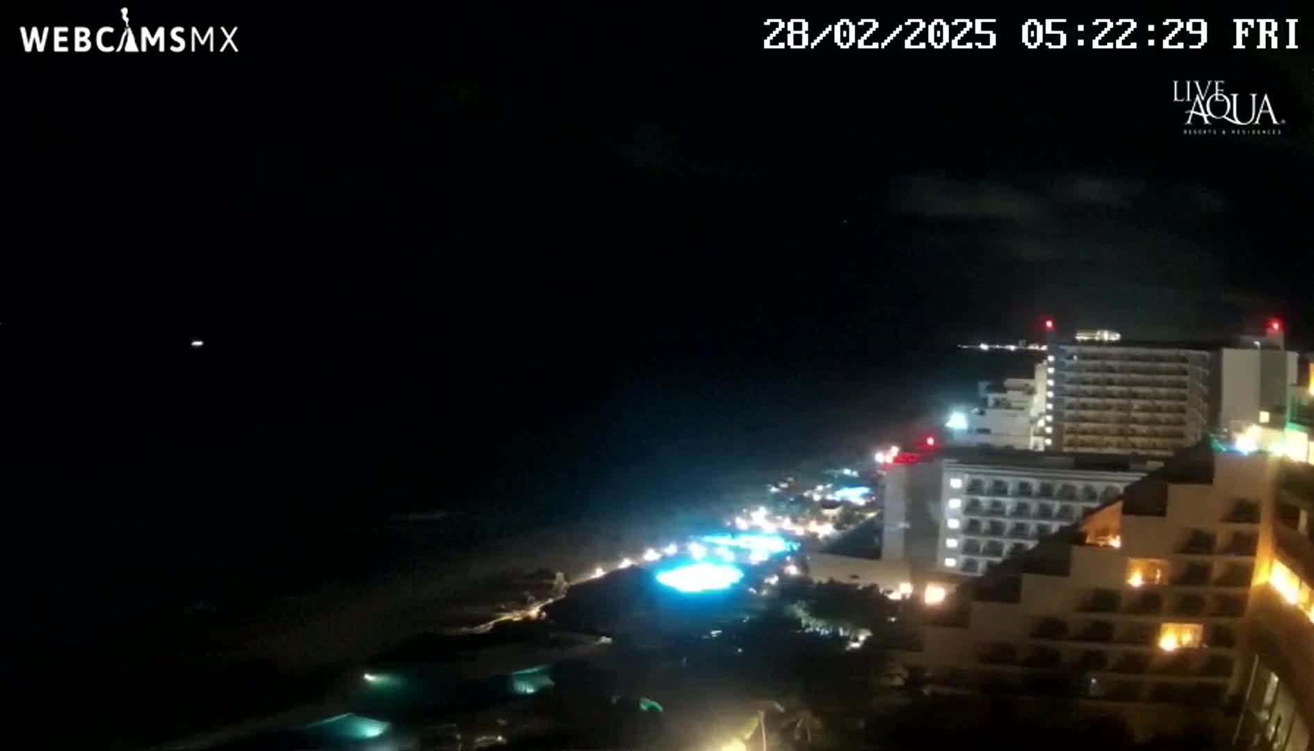 A nighttime coastal view shows a dark ocean, a lit beach, and several brightly lit high-rise hotels under a mostly clear, dark sky.