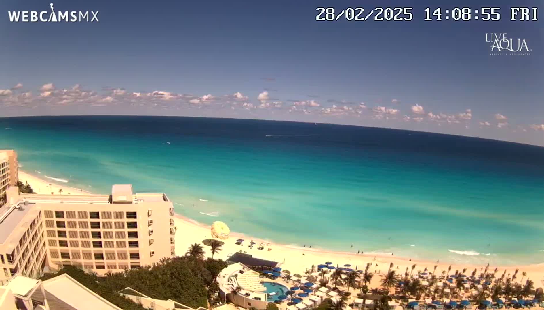 A high-angle view shows a beach with turquoise water under a mostly sunny sky, and a large hotel partially visible in the foreground.