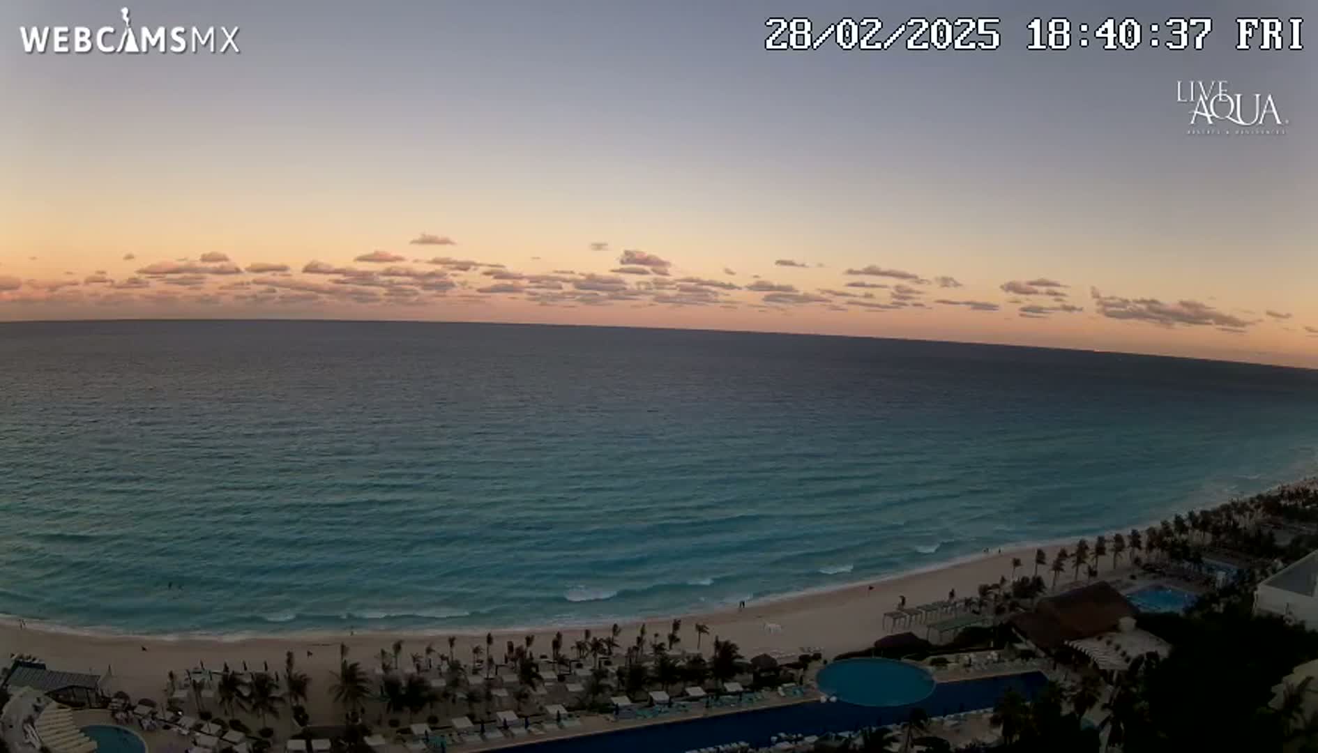 A tranquil ocean scene at sunset, with calm waves lapping a sandy beach lined with palm trees and a resort complex.